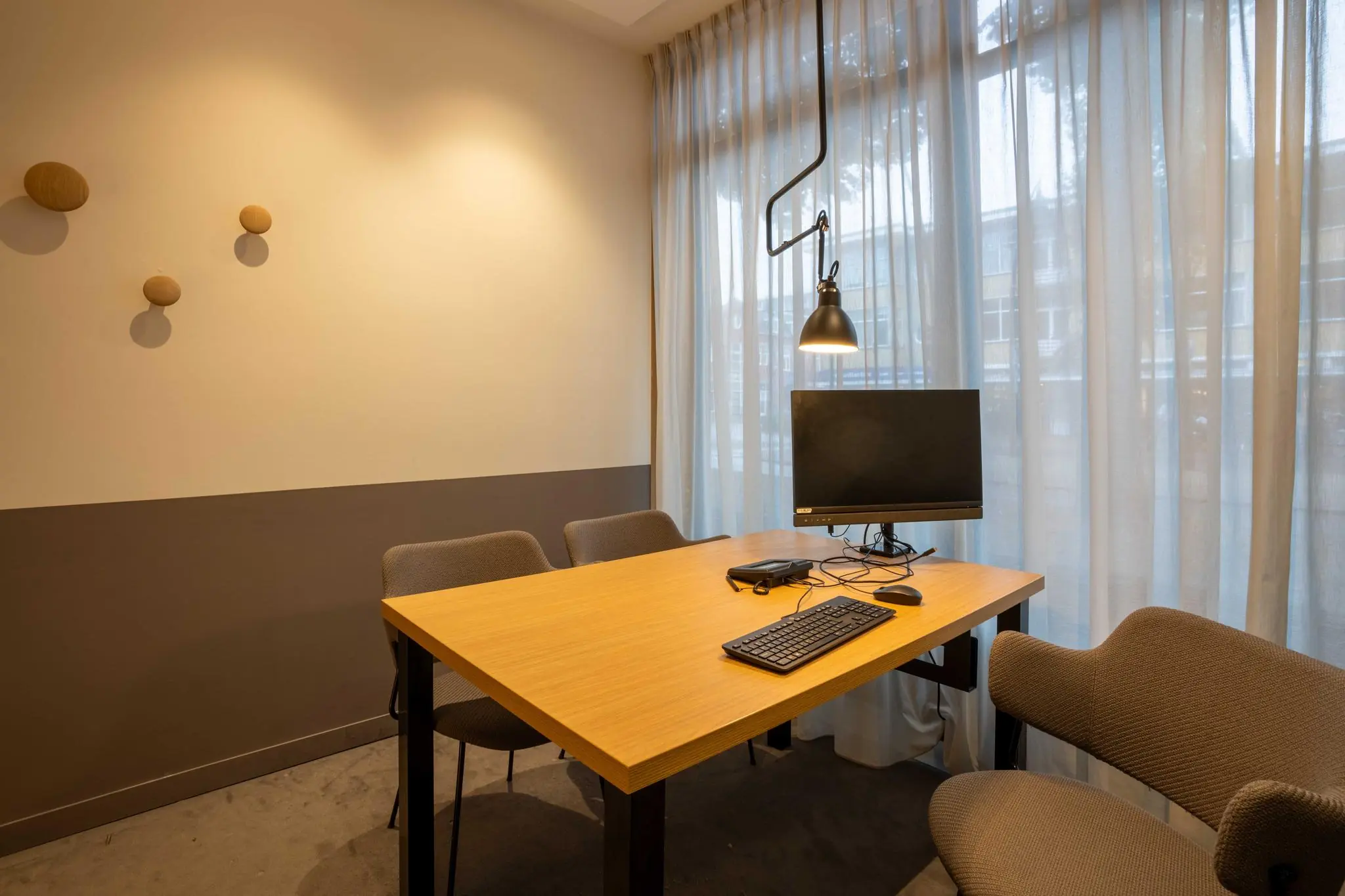 Kleine vergaderruimte aan de 1e Hogeweg met houten tafel, vier stoelen, computerscherm en hanglamp bij een raam met lichte gordijnen.